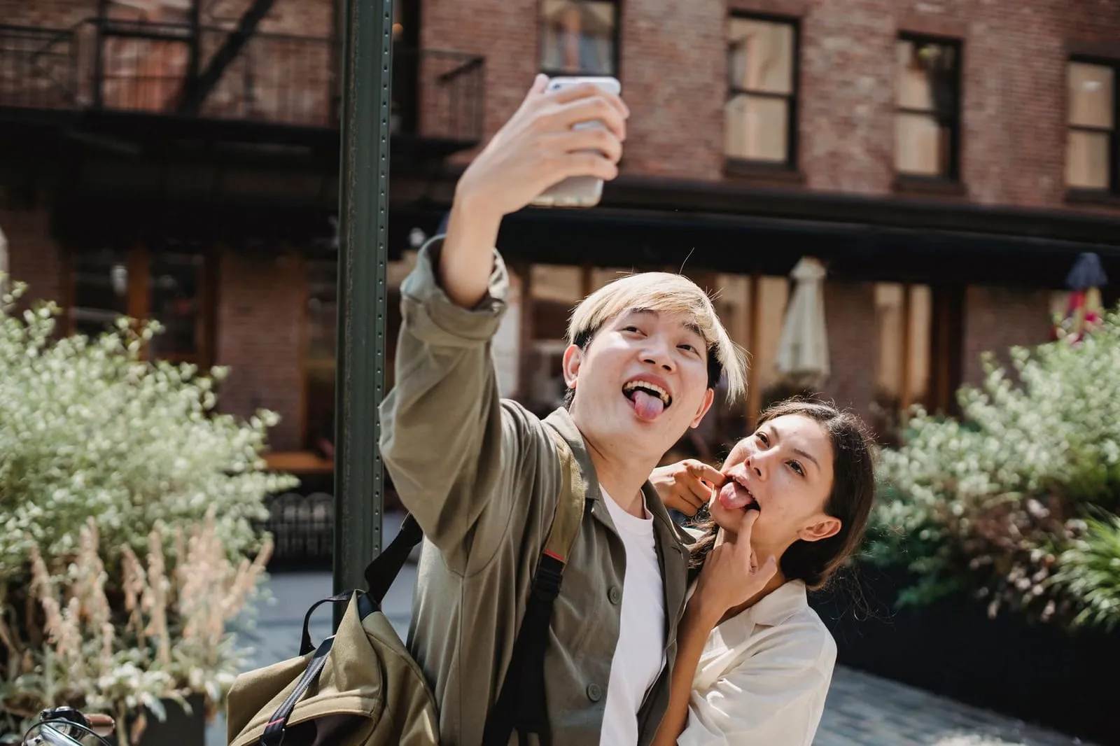 Playful young Asian couple showing tongue and taking selfie on smartphone while having fun on street on sunny day