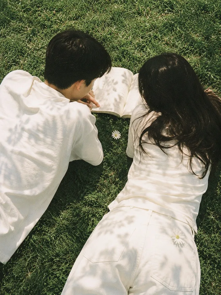 A couple lying side by side on the grass, reading a book together in dappled sunlight