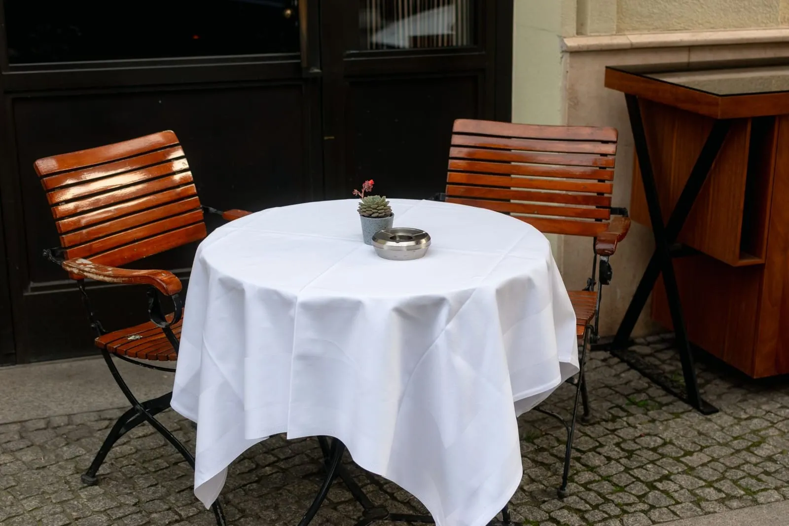 Cozy café setting with wooden chairs and a white tablecloth on a cobblestone street.