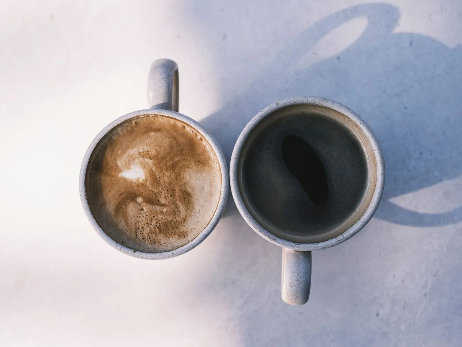 Two coffee cups side by side on a sunlit table, one a latte and one black coffee, casting long shadows.